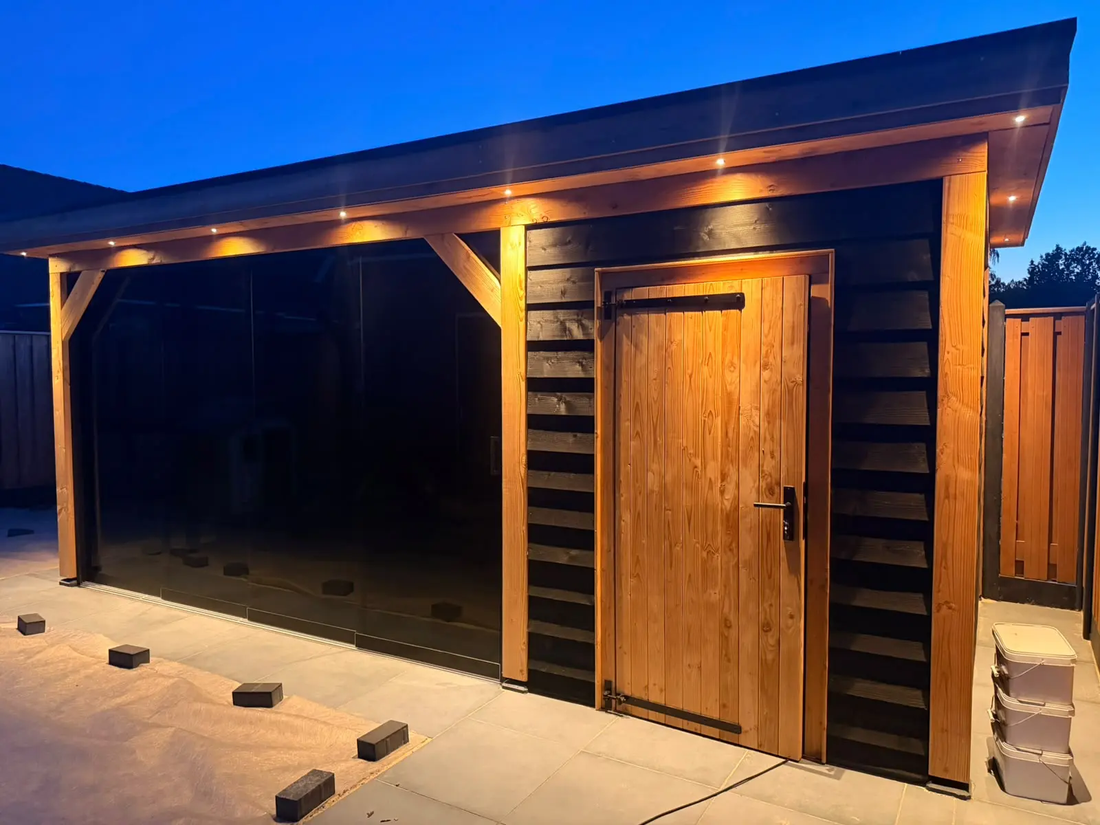 Houten overkapping in de avond met verlichting en glazen schuifwand